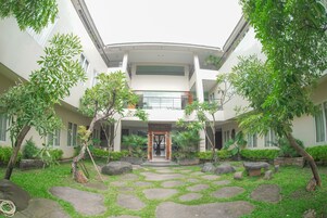 A courtyard with a central stone pathway, surrounded by greenery and trees, leading to a building entrance with a glass door.