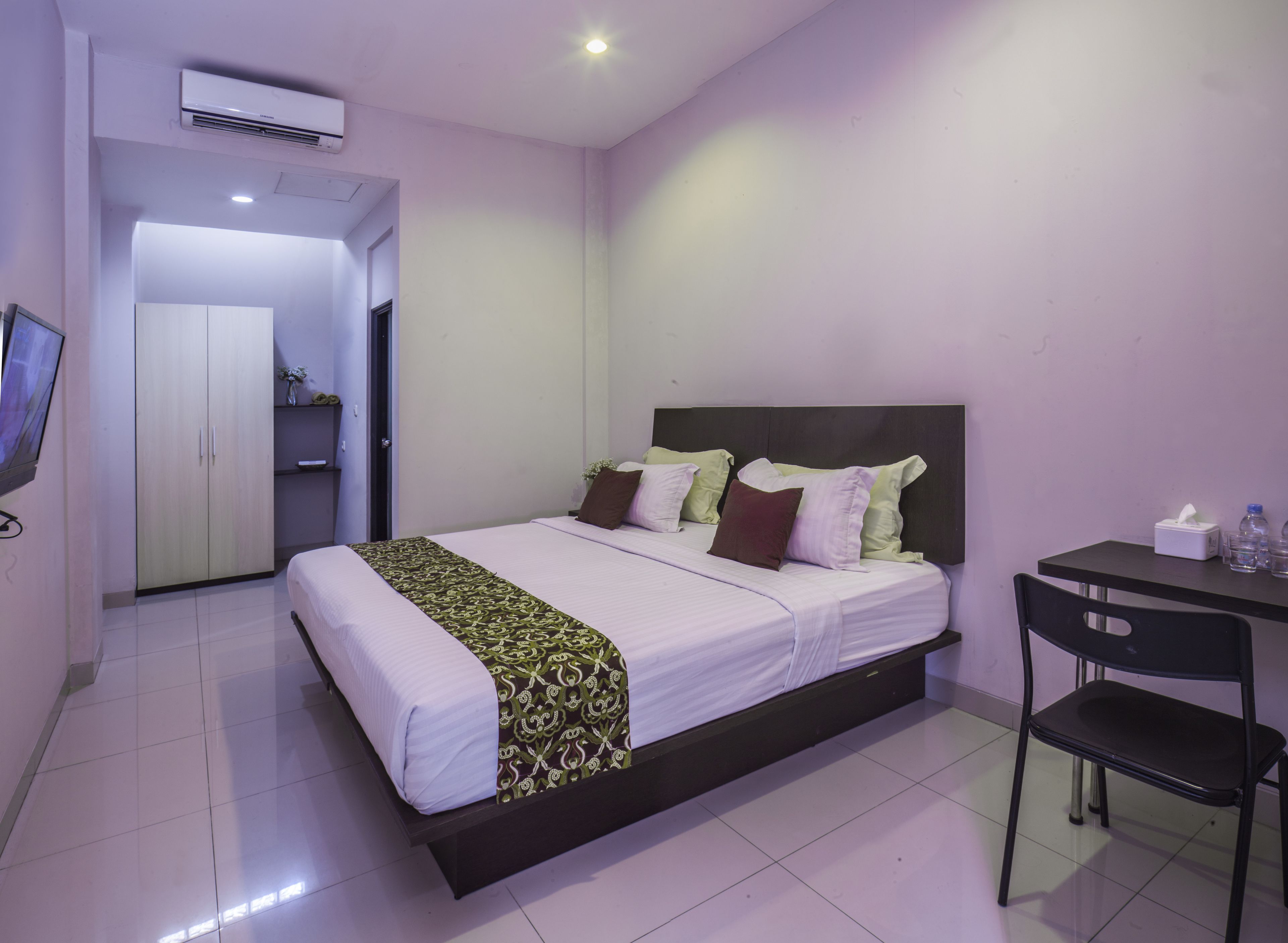 A bedroom with a bed, a desk, and a chair. There is a wardrobe and an air conditioning unit.