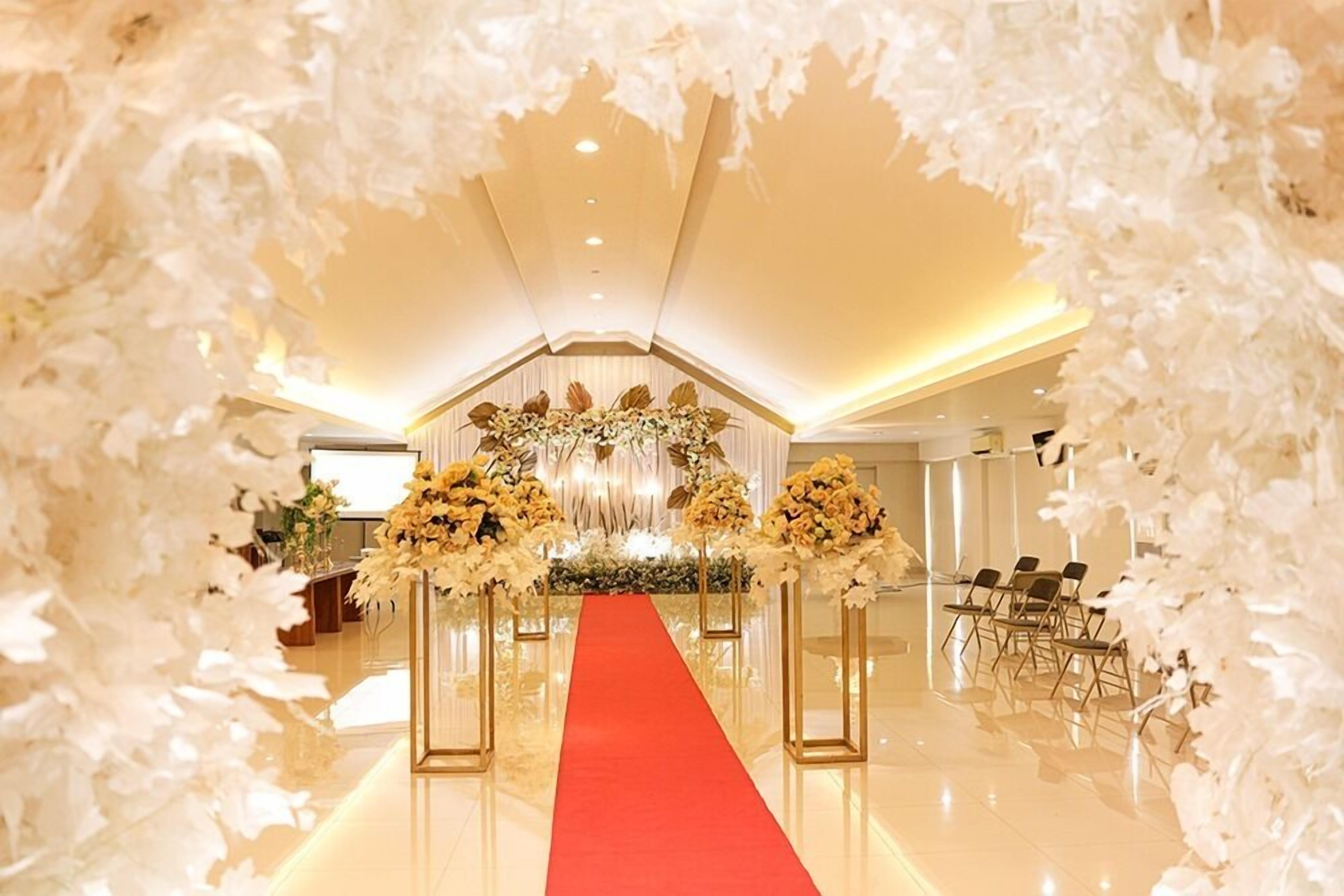 Un hall de hotel con una alfombra roja que conduce a una recepción, flanqueado por arreglos florales blancos y un candelabro.