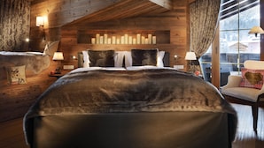 A cozy bedroom with a wooden ceiling, a large bed with a fur throw, a wall-mounted shelf with candles, and a view of a snowy landscape through a window. - Hotel Arlberg (Lech am Arlberg)