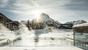 A snow-covered outdoor pool with steam rising, surrounded by buildings and mountains under a clear sky. - Hotel Arlberg (Lech am Arlberg)