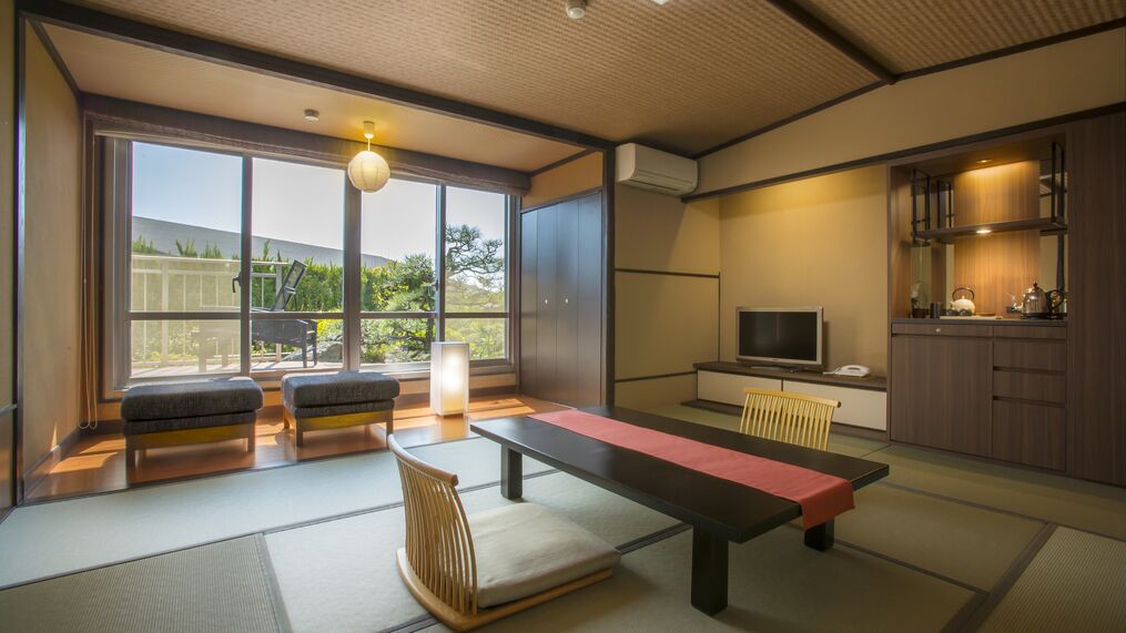 A traditional Japanese room with tatami flooring, a low table, cushions, a television, and a kitchen area.
