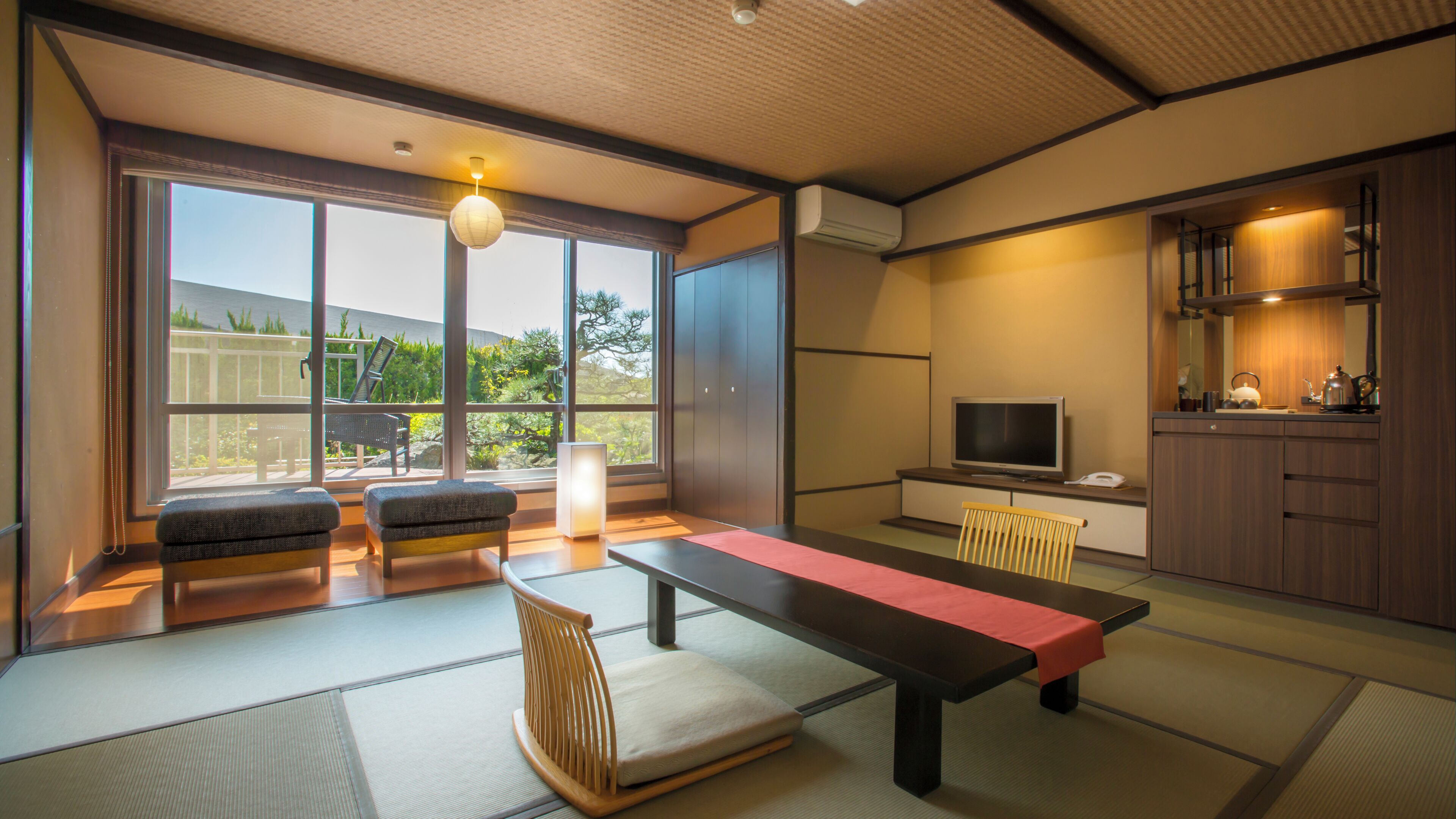 Una habitación tradicional japonesa con suelo de tatami, una mesa baja, asientos, un televisor y vista a un jardín.