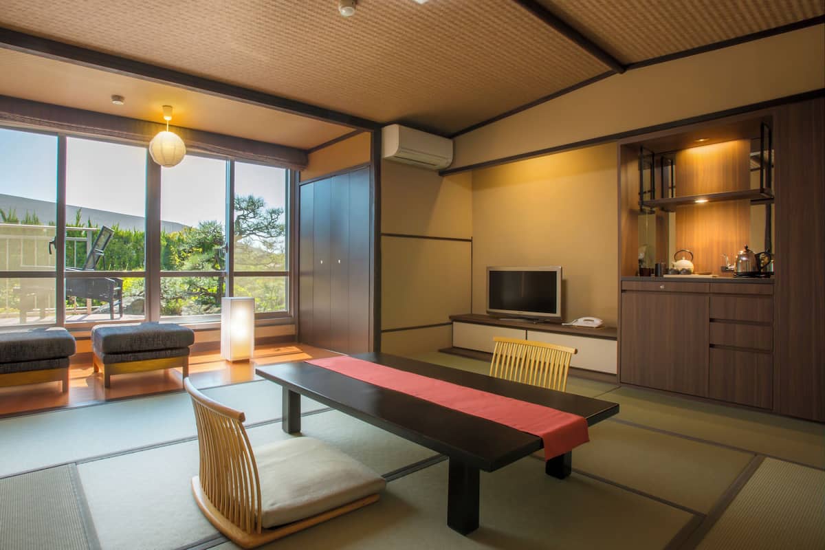 A traditional Japanese-style room with tatami flooring, a low table, seating, a television, and a view of a garden.