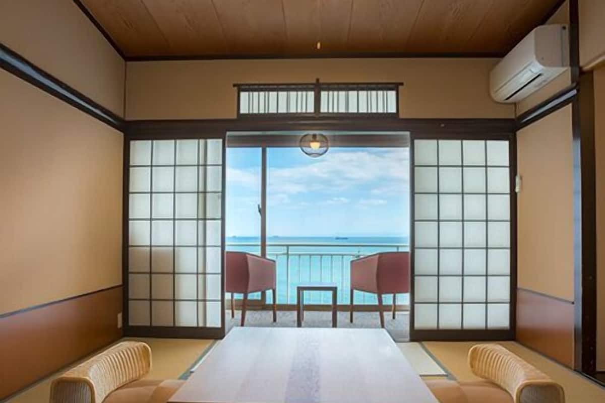 A traditional Japanese room with tatami flooring, a low table, and sliding doors leading to a balcony with a view of the sea.