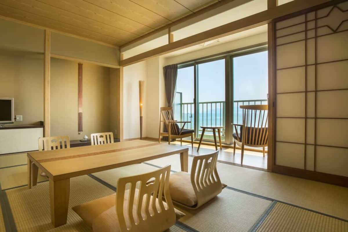 A traditional Japanese room with tatami flooring, a low table, chairs, a TV, and a sliding door leading to a balcony.