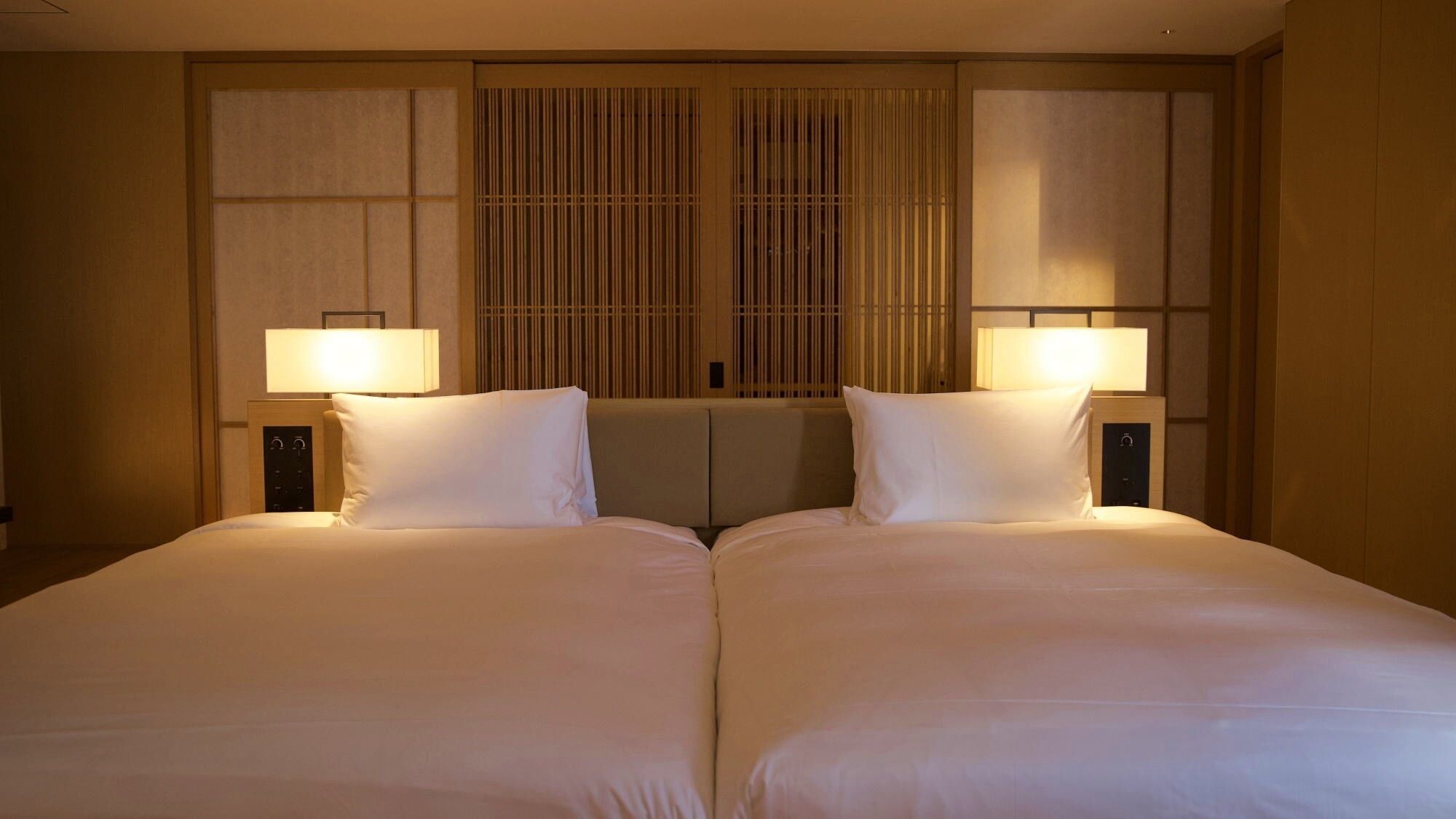 Habitación de hotel con dos camas, un ventanal grande con persianas verticales y lámparas de noche.