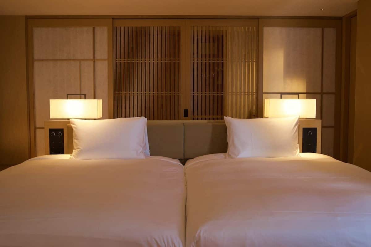 A hotel room with two beds, a large window with vertical blinds, and bedside lamps.