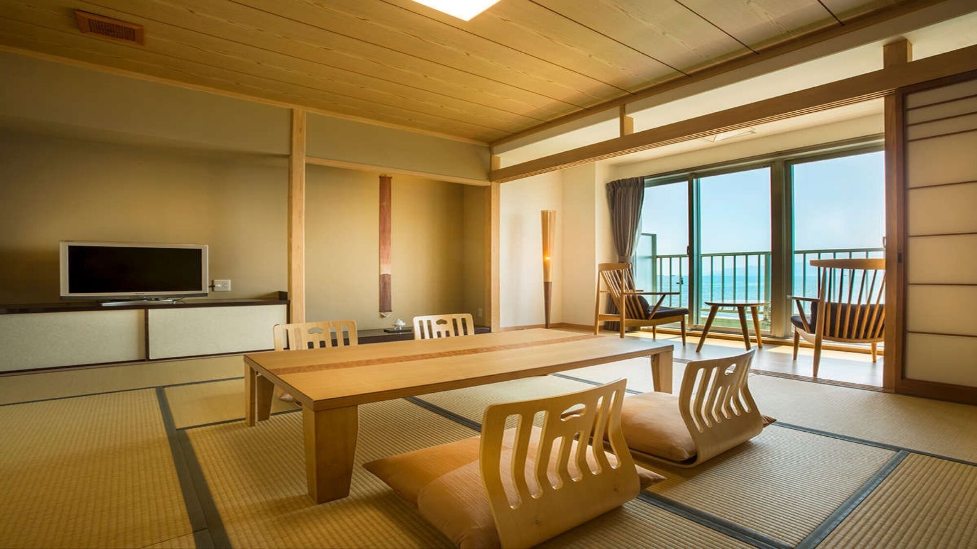Una habitación japonesa tradicional con suelo de tatami, una mesa baja, sillas, televisión y un balcón con vistas.
