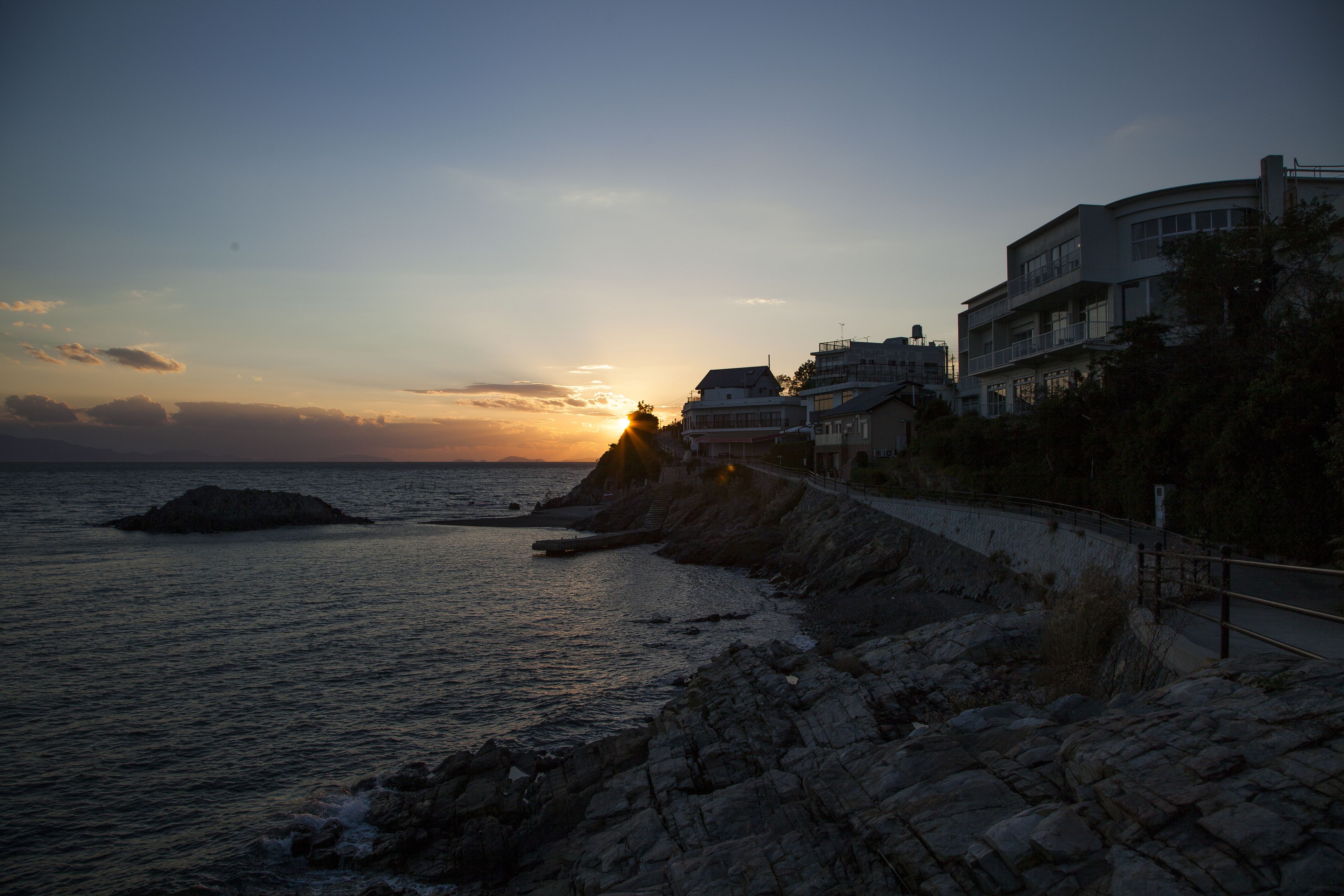 A coastal scene with a sunset, rocky shoreline, and buildings along the edge.