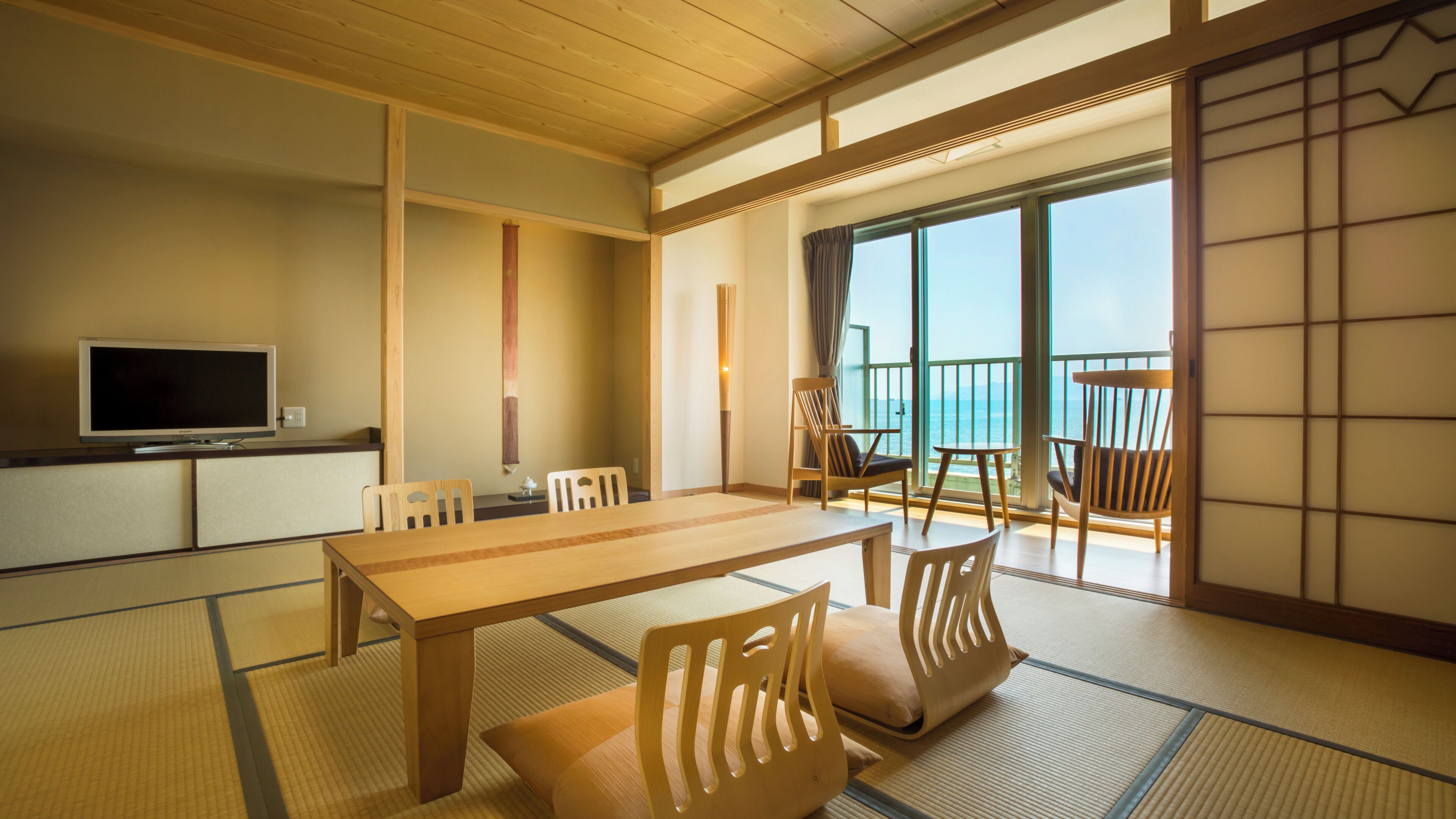 Una habitación japonesa tradicional con suelo de tatami, una mesa baja, sillas, un televisor y vista al mar.