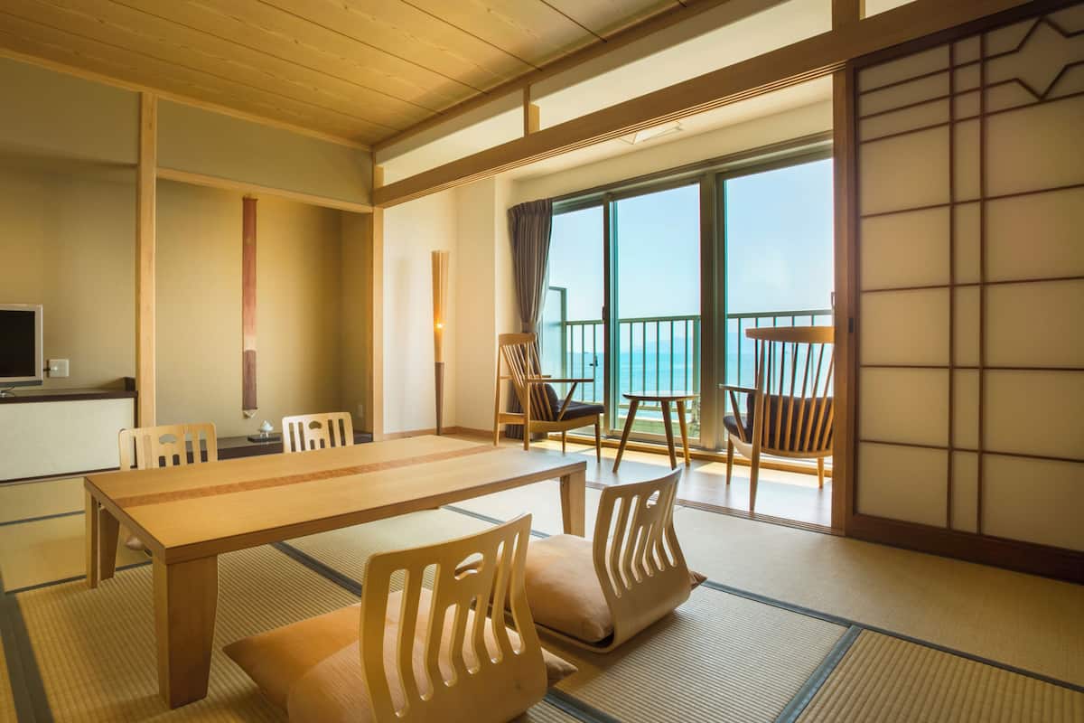 A traditional Japanese room with tatami flooring, a low table, chairs, a television, and a view of the sea.
