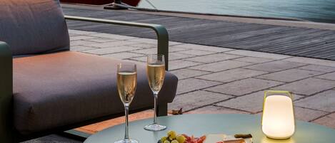 Una mesa con una copa de vino, una botella de champán y una tabla de embutidos y quesos. Un barco está amarrado al fondo, y edificios bordean el paseo marítimo.