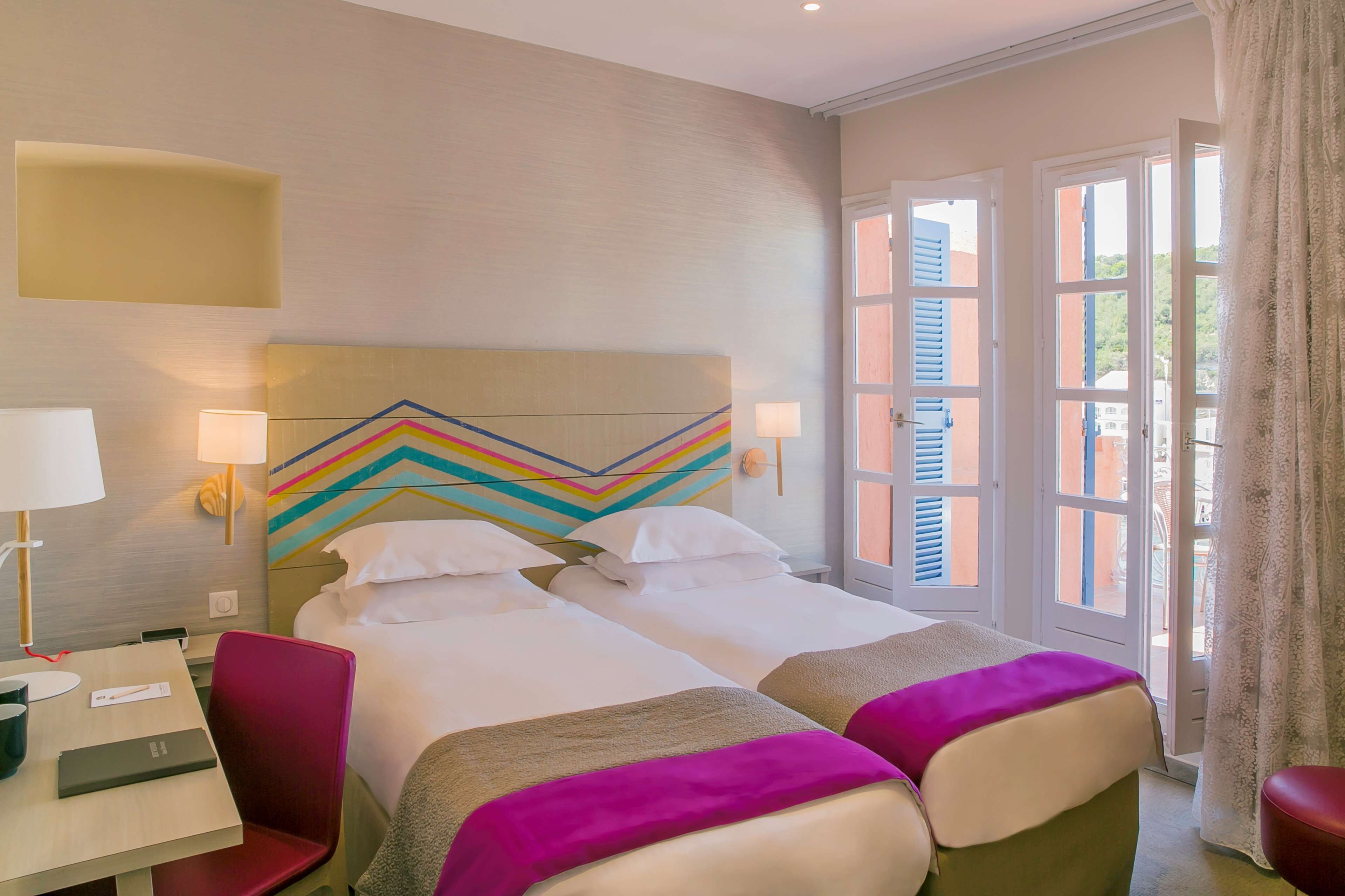 A hotel room with two beds, a desk, and a chair. There are two bedside tables with lamps. The room has a large window with shutters and a view of buildings outside.