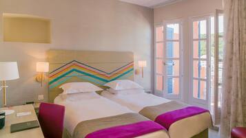 Quarto de hotel com duas camas, uma escrivaninha e uma cadeira. Há duas mesas de cabeceira com abajures. O quarto tem uma janela grande com persianas e vista para edifícios.