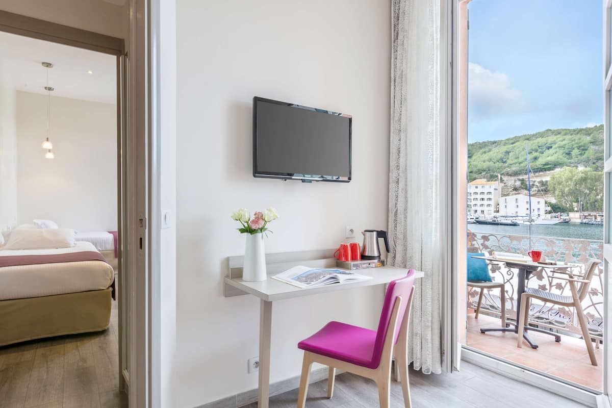 Une chambre d’hôtel comprenant un lit, un bureau avec une chaise, un téléviseur à écran plat, un vase avec des fleurs et une vue sur un balcon aménagé avec du mobilier d’extérieur.