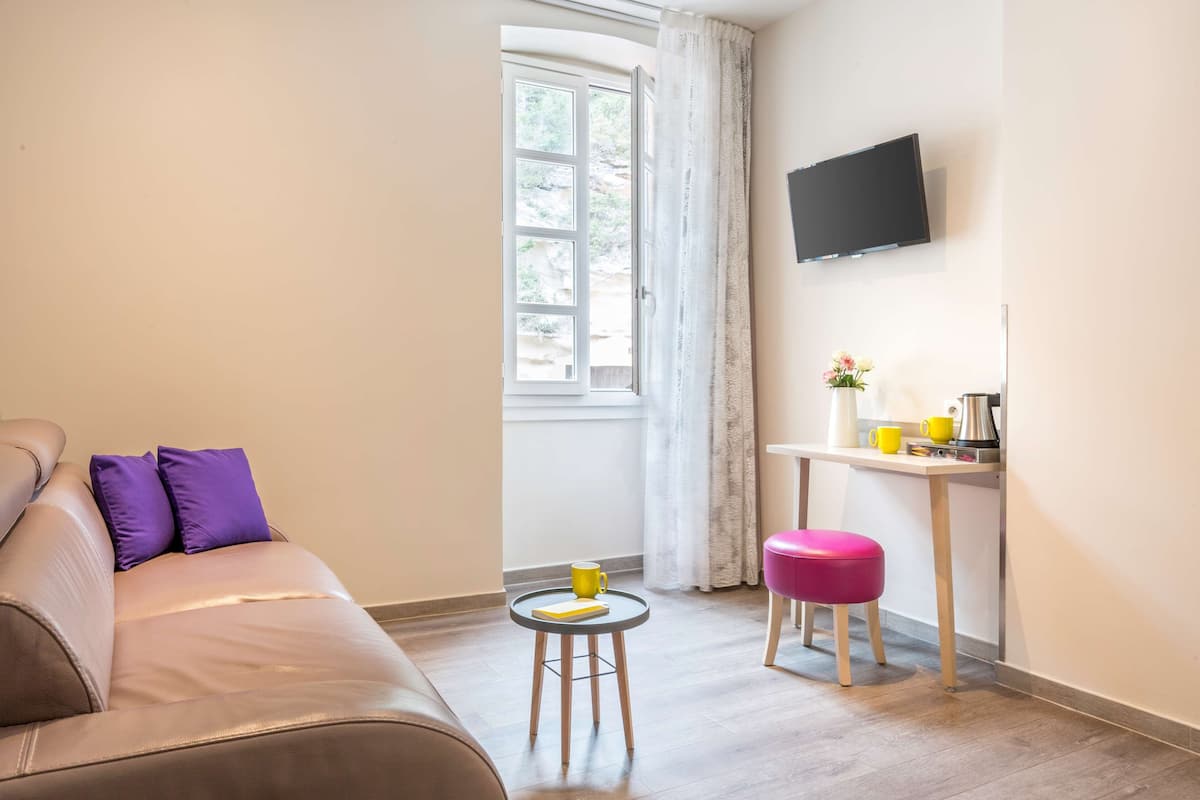 Un salon moderne avec un canapé, une petite table ronde sur laquelle se trouve une tasse jaune, un tabouret rose, un téléviseur à écran plat fixé au mur et une fenêtre avec des rideaux.