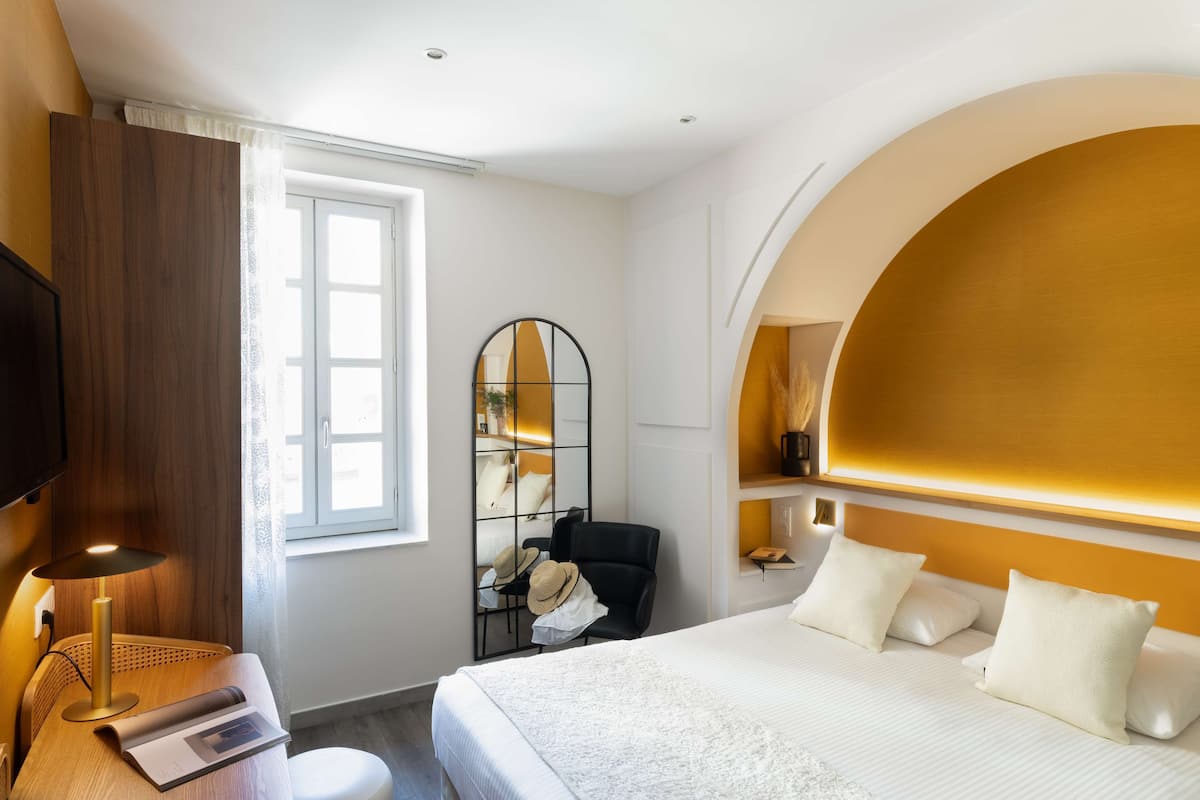 Une chambre d’hôtel avec un grand lit, un bureau avec une lampe, une chaise, un miroir et une fenêtre.