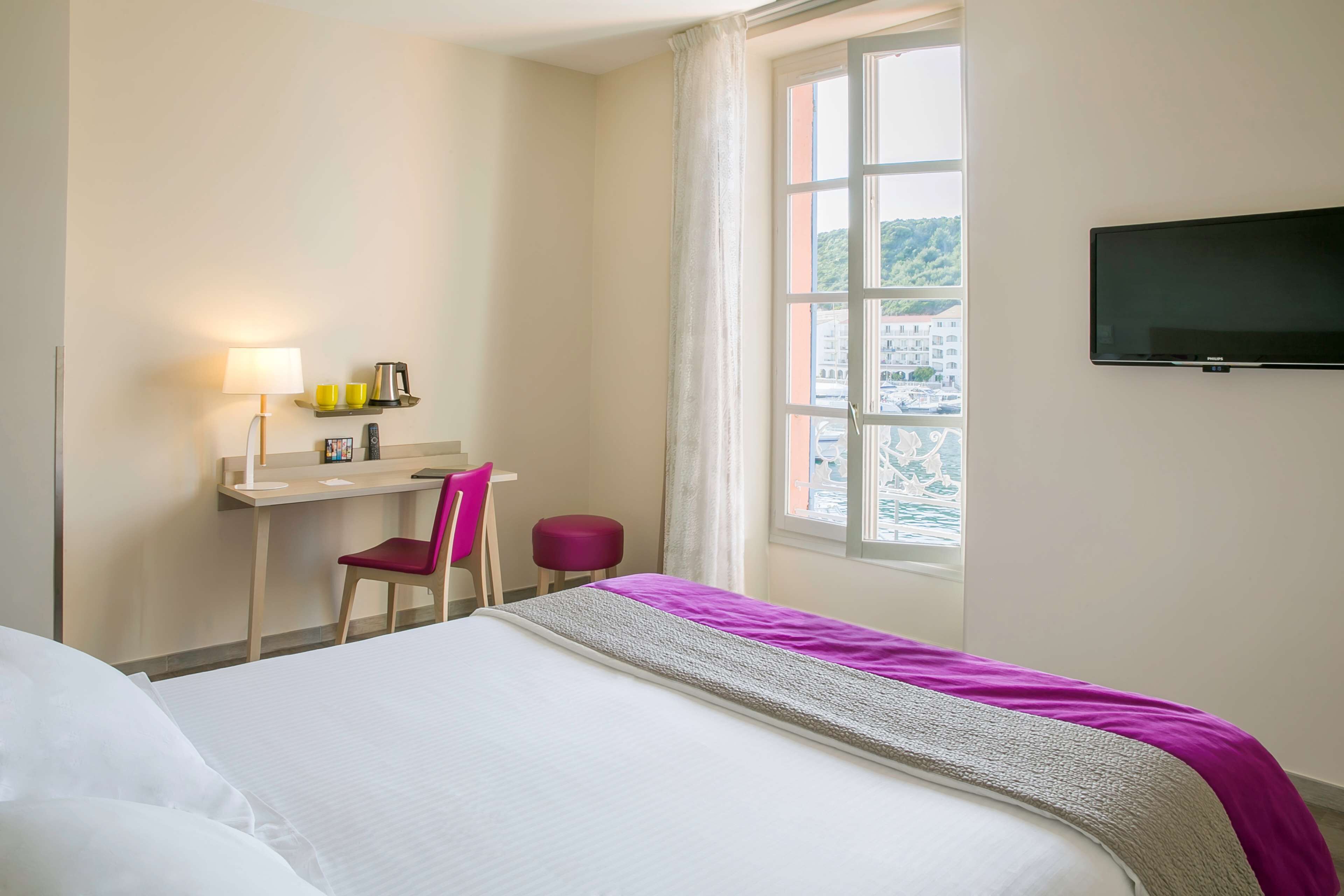 A hotel room with a bed, a desk with a chair, a TV, and a window with a view.