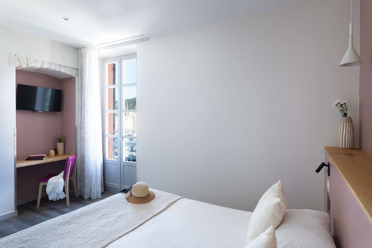 Une chambre à coucher comprenant un lit, un bureau avec une chaise, un téléviseur fixé au mur et une fenêtre avec des rideaux.