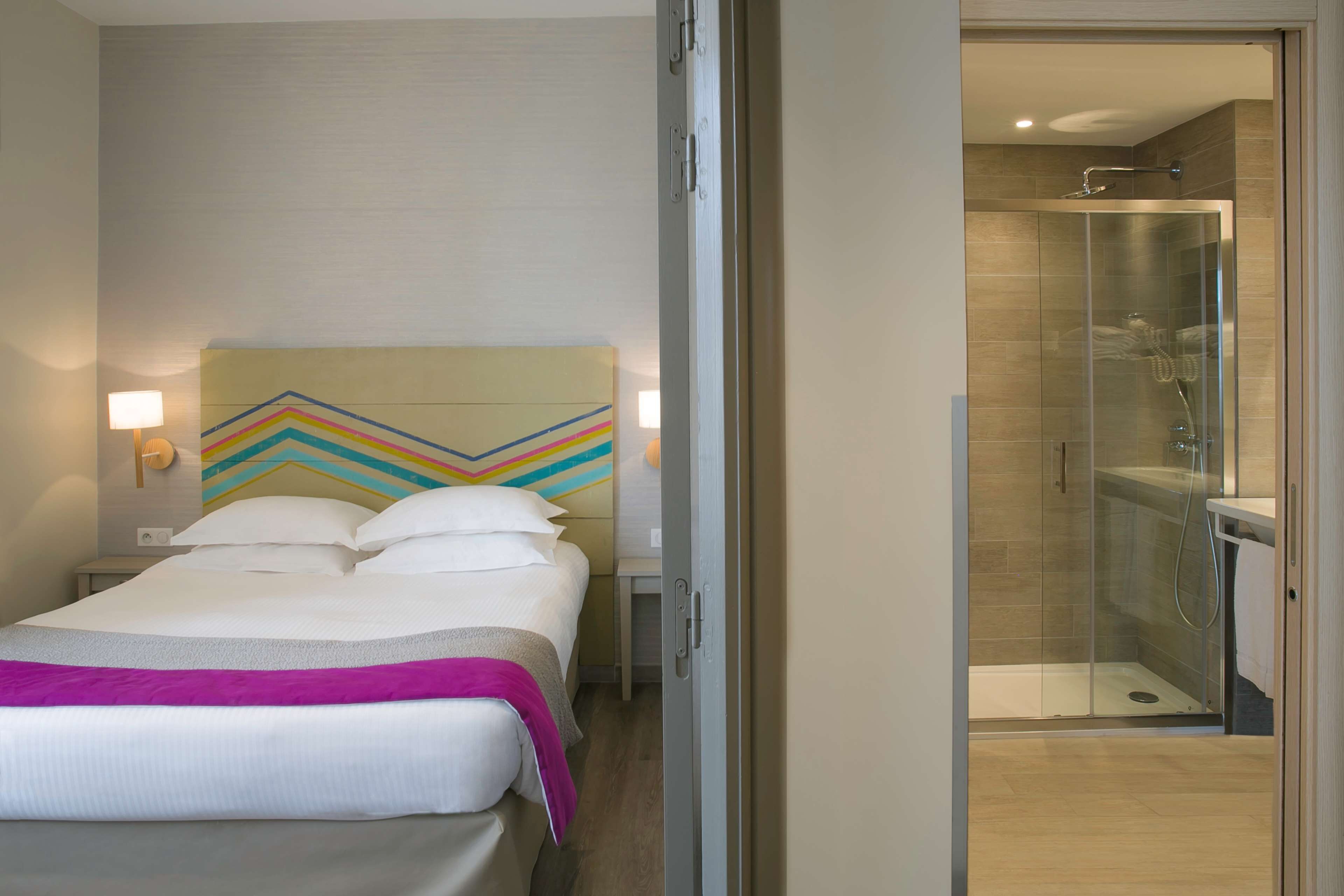A hotel room with a bed, a nightstand, a wall with a colorful geometric design, and a glass-enclosed shower area.