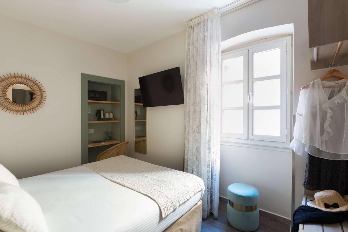 Une chambre à coucher comprenant un lit, un téléviseur fixé au mur, une armoire, une fenêtre avec des rideaux et un petit tabouret.