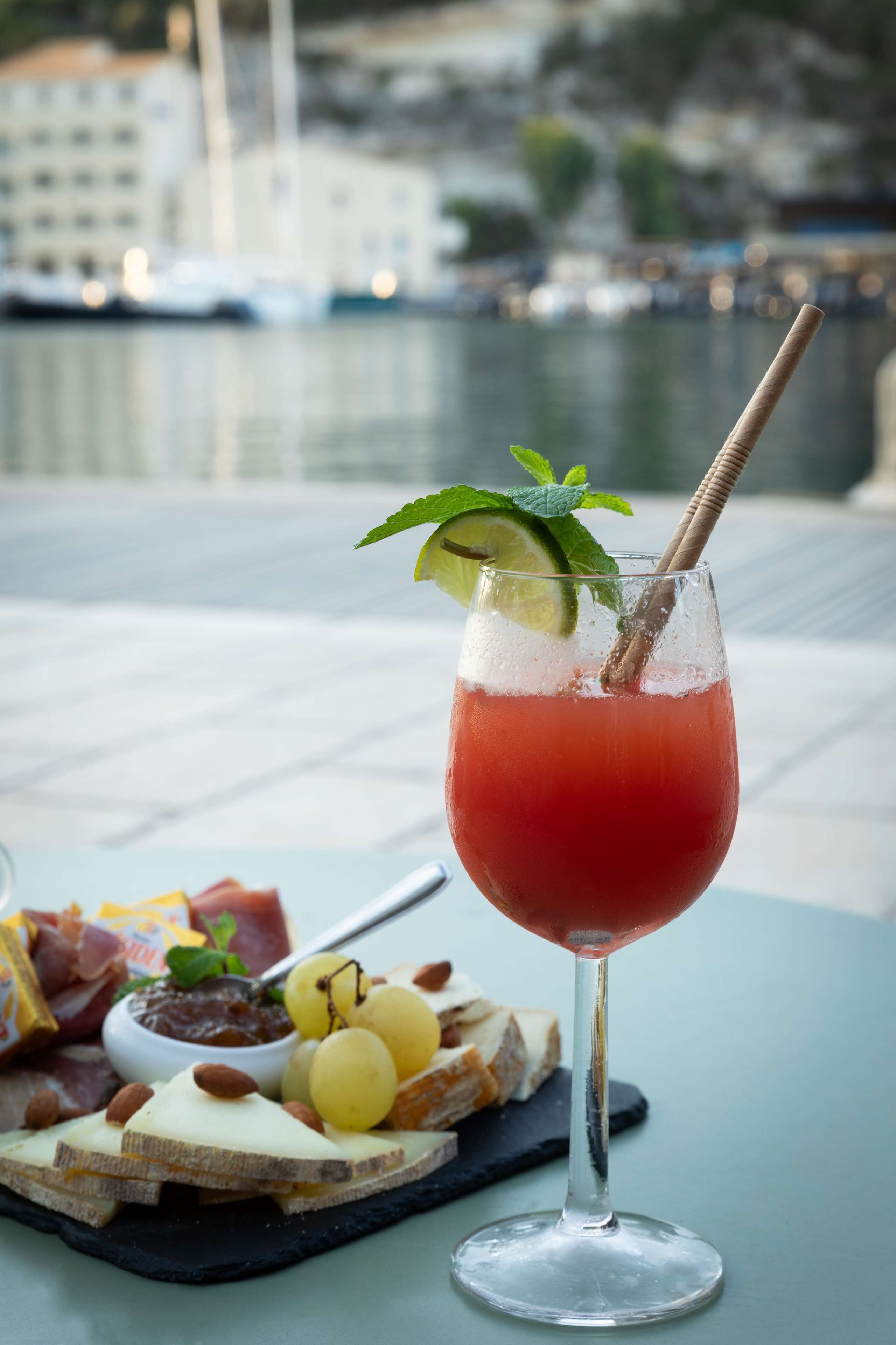 A table with a glass of red drink garnished with mint and lime, a cheese board with various cheeses and nuts, and a small bowl of jam.