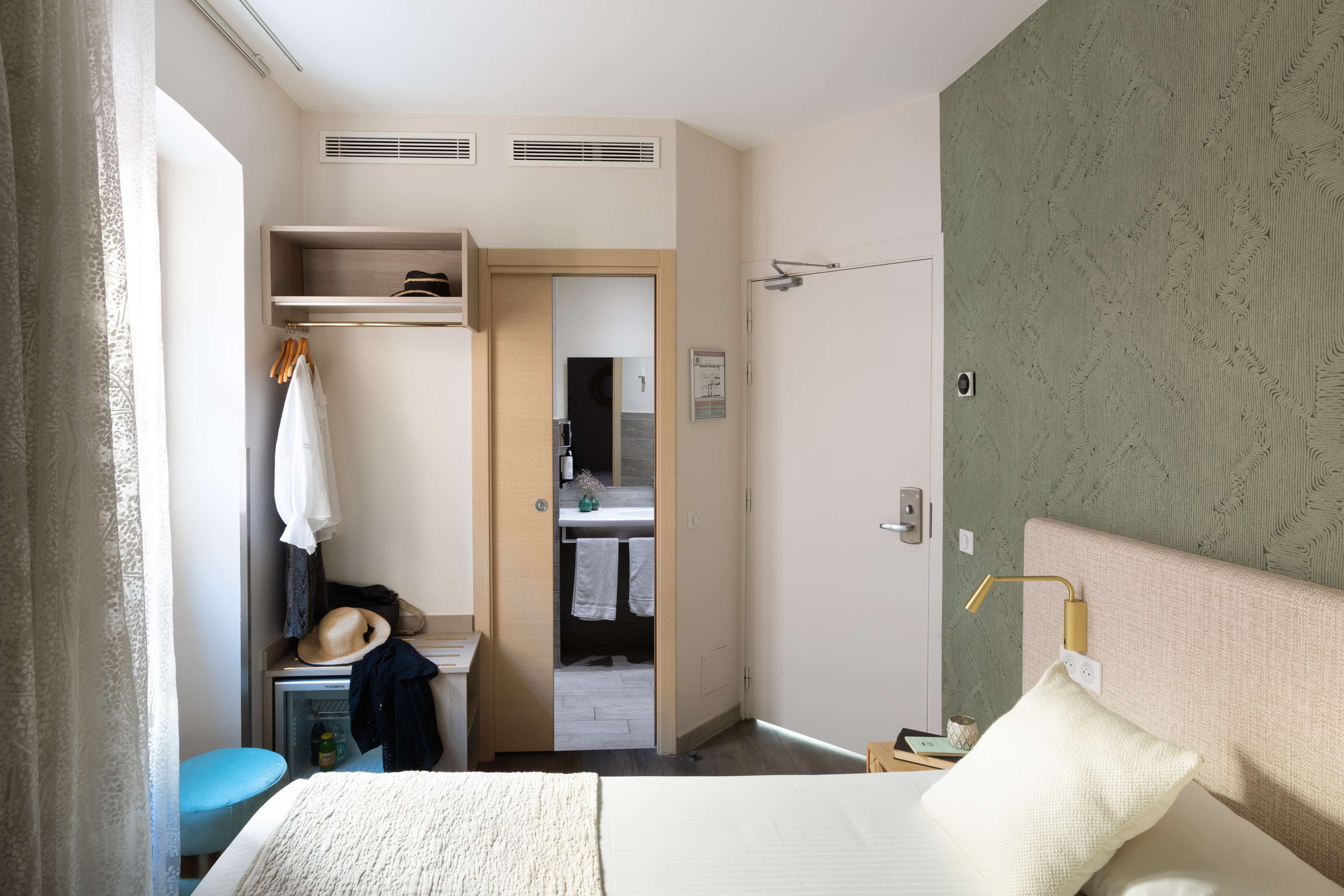 Une chambre d’hôtel avec un lit, une armoire, un miroir et une salle de bain visible à travers une porte ouverte.