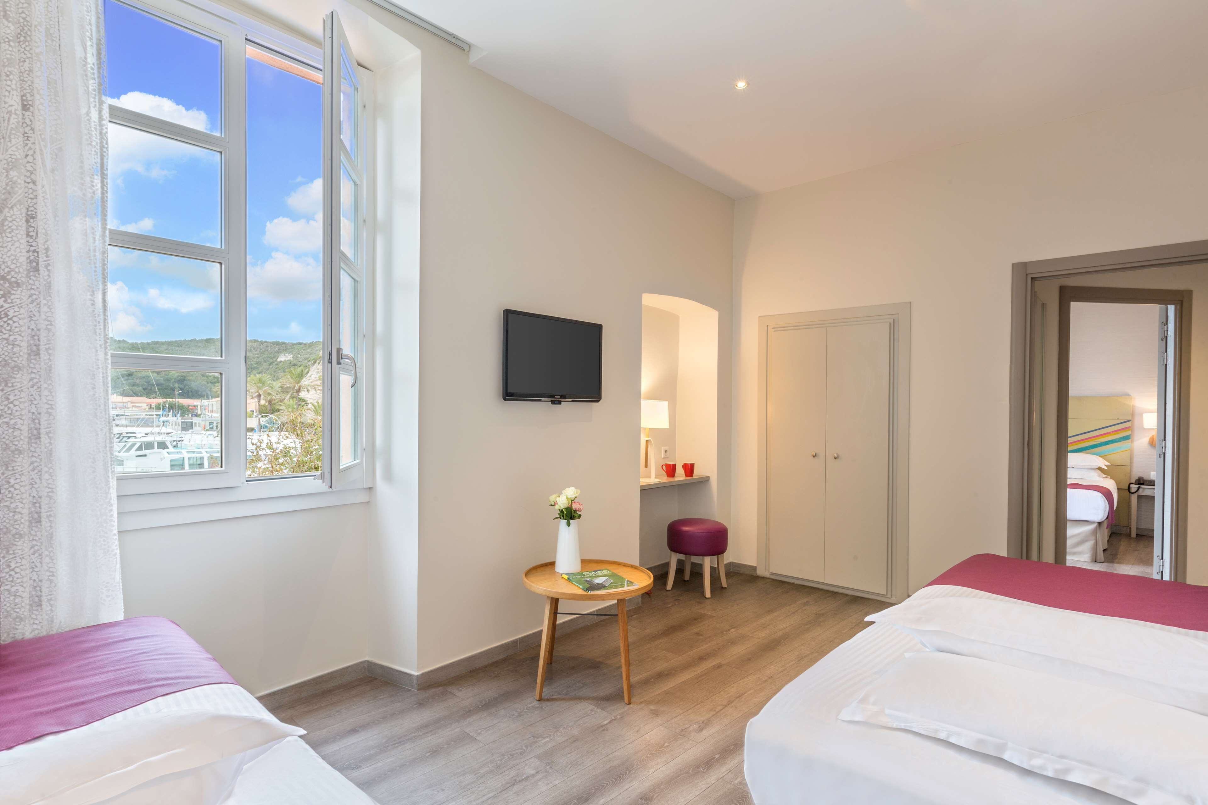 A hotel room with two beds, a TV, a small table with a vase, and a view of the city through a large window.