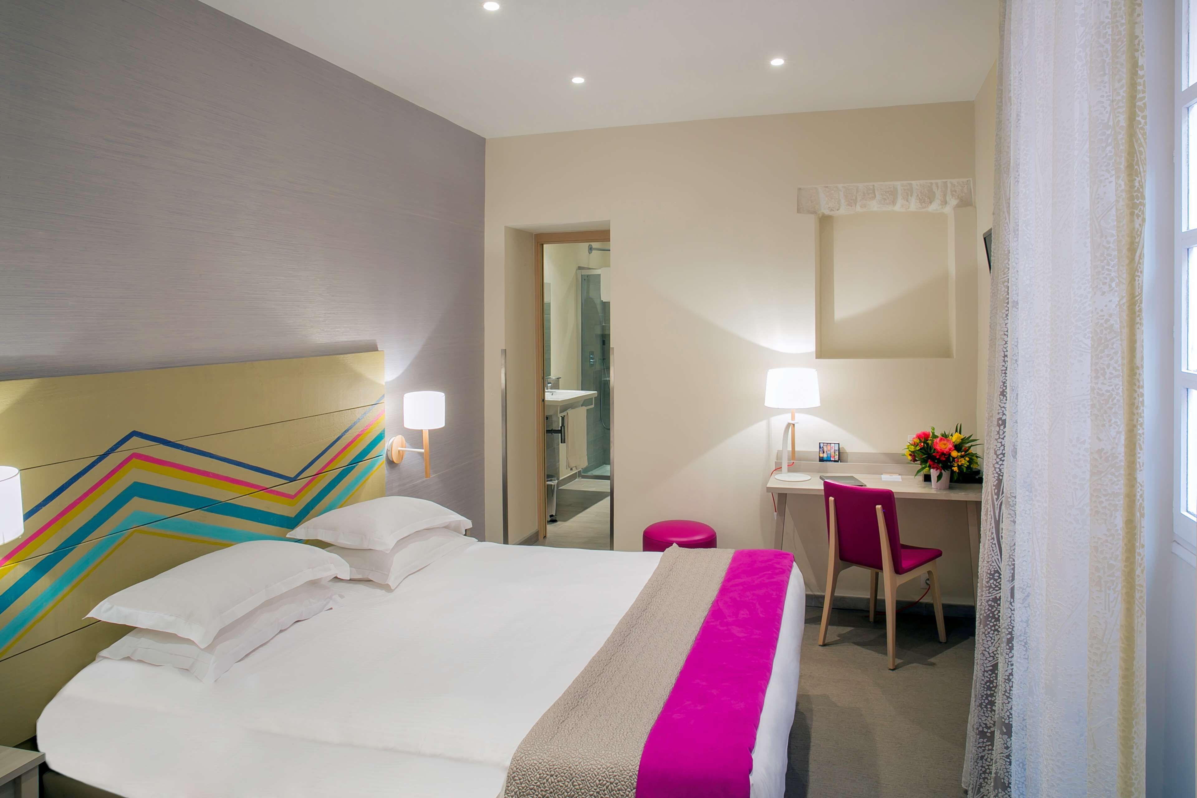 Habitación de hotel con cama, escritorio, silla, espejo y ventana con cortinas.