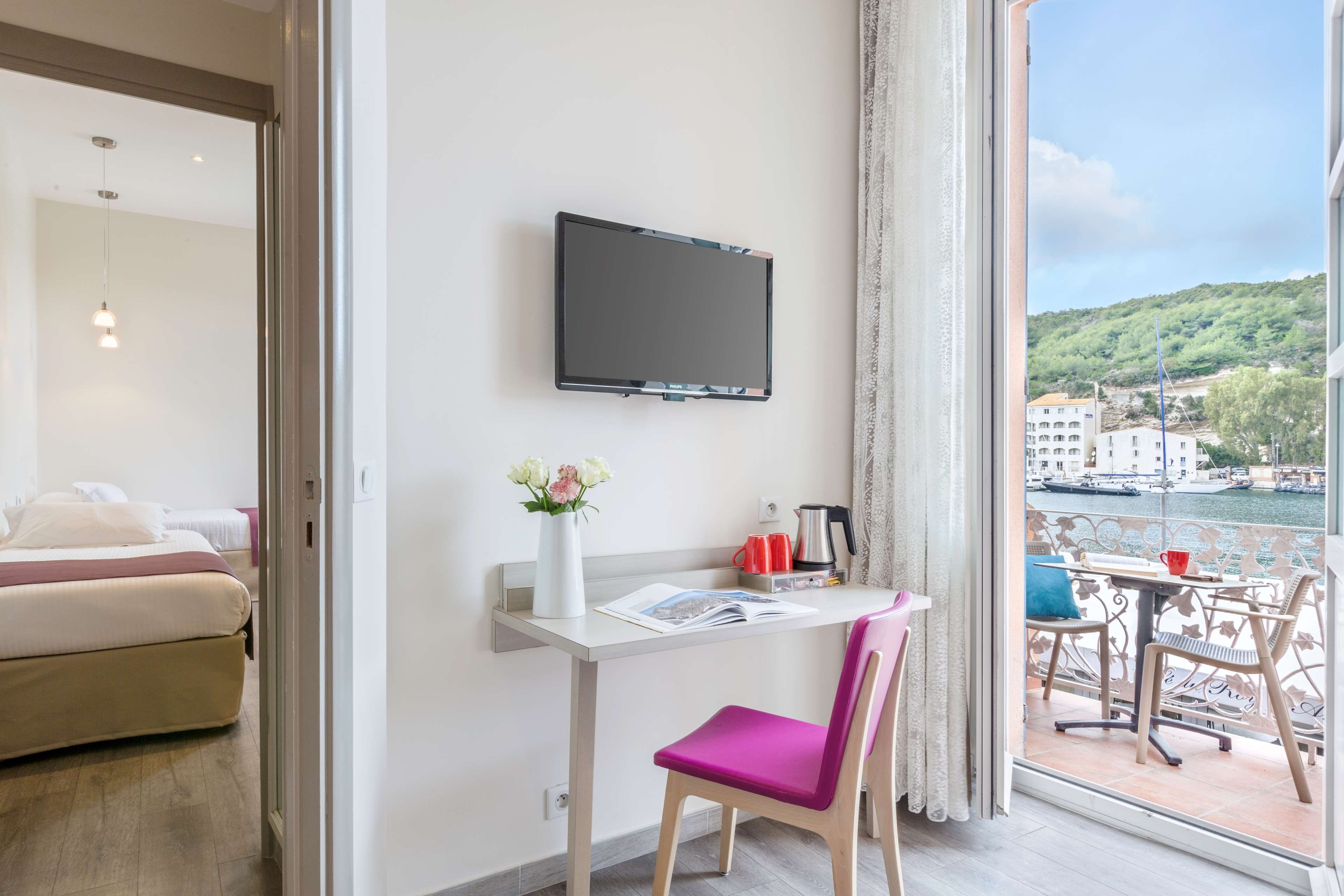 Habitación de hotel con una cama, un escritorio con una silla, un televisor de pantalla plana, un jarrón con flores y vistas a un cuerpo de agua con edificios.