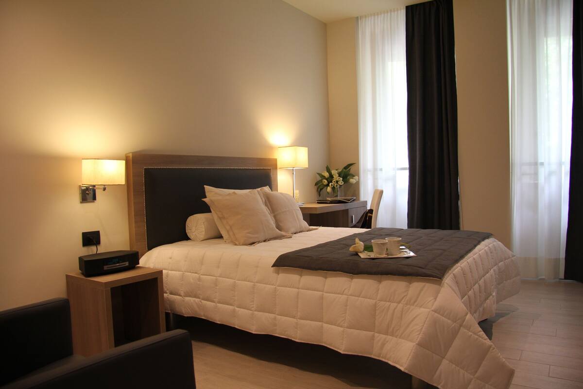 A hotel room with a large bed, bedside lamps, a small table with a plant, and a view of the outside through the window.