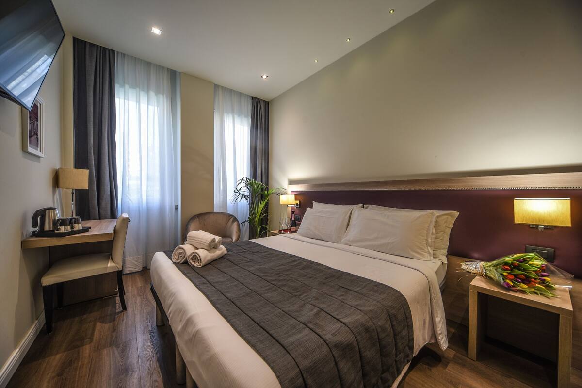 A hotel room with a bed, a desk, a chair, a nightstand with a lamp, and a bouquet of flowers.