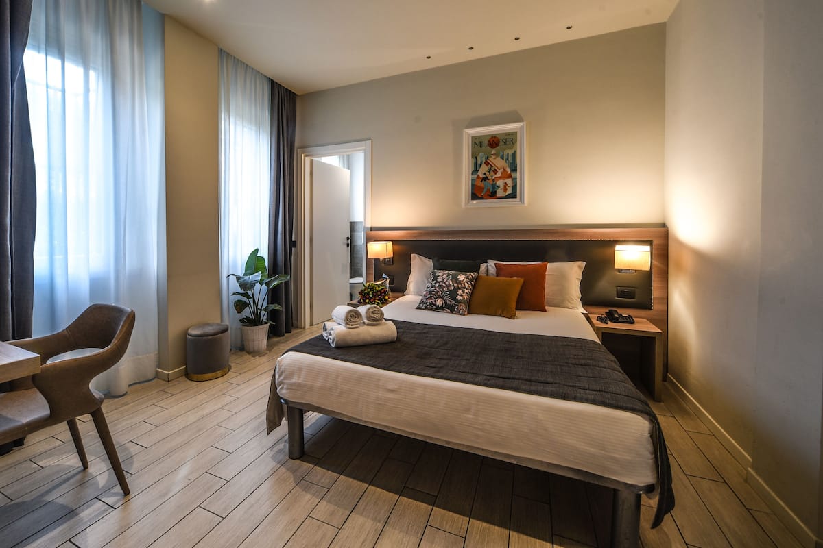 A hotel room with a large bed, a desk, a chair, a potted plant, and a painting on the wall.