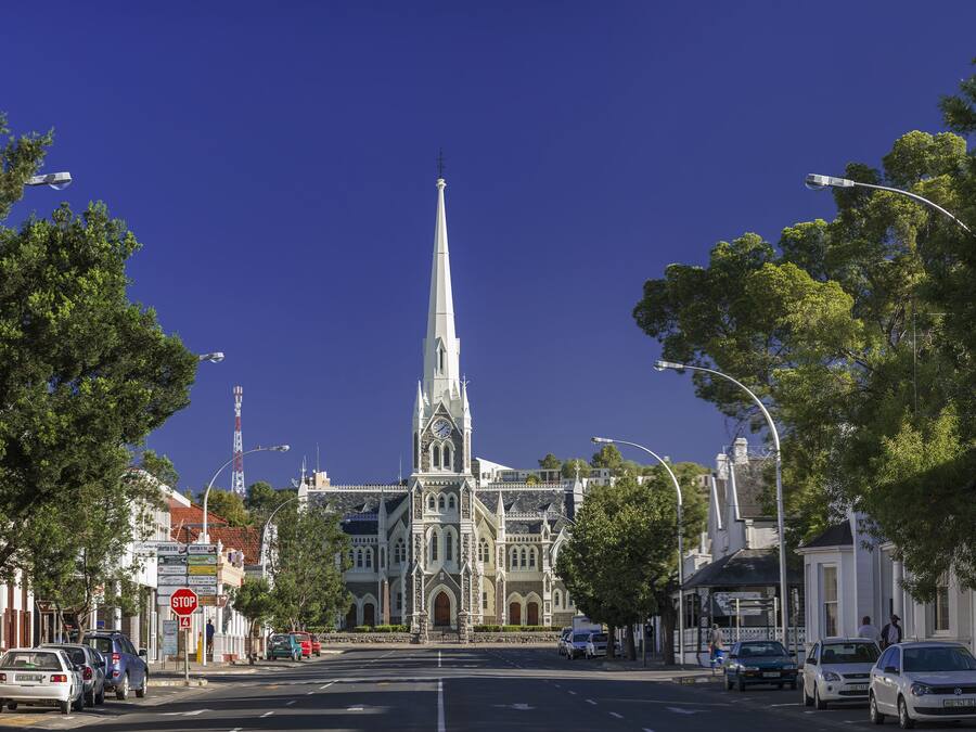 Vista de una calle con una imponente aguja de iglesia, coches estacionados y un cielo azul despejado.