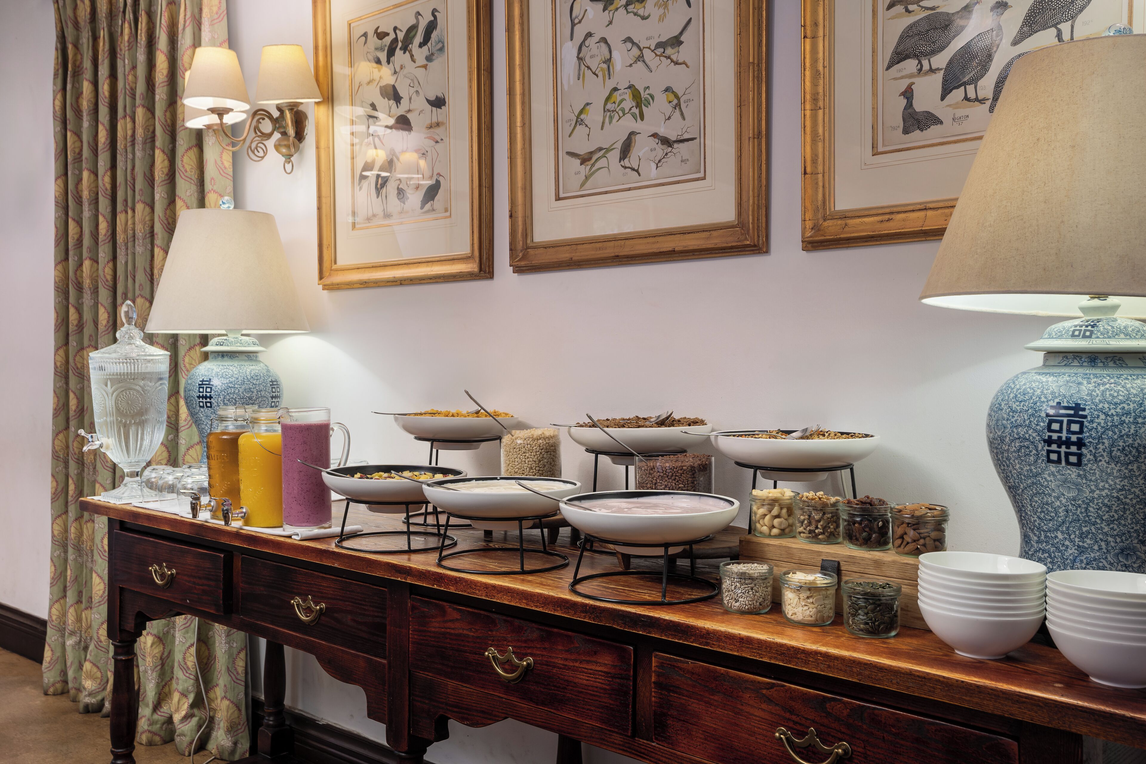 Un coin repas avec une table en bois, dressée avec différents plats, une lampe et des illustrations d’oiseaux encadrées accrochées au mur.