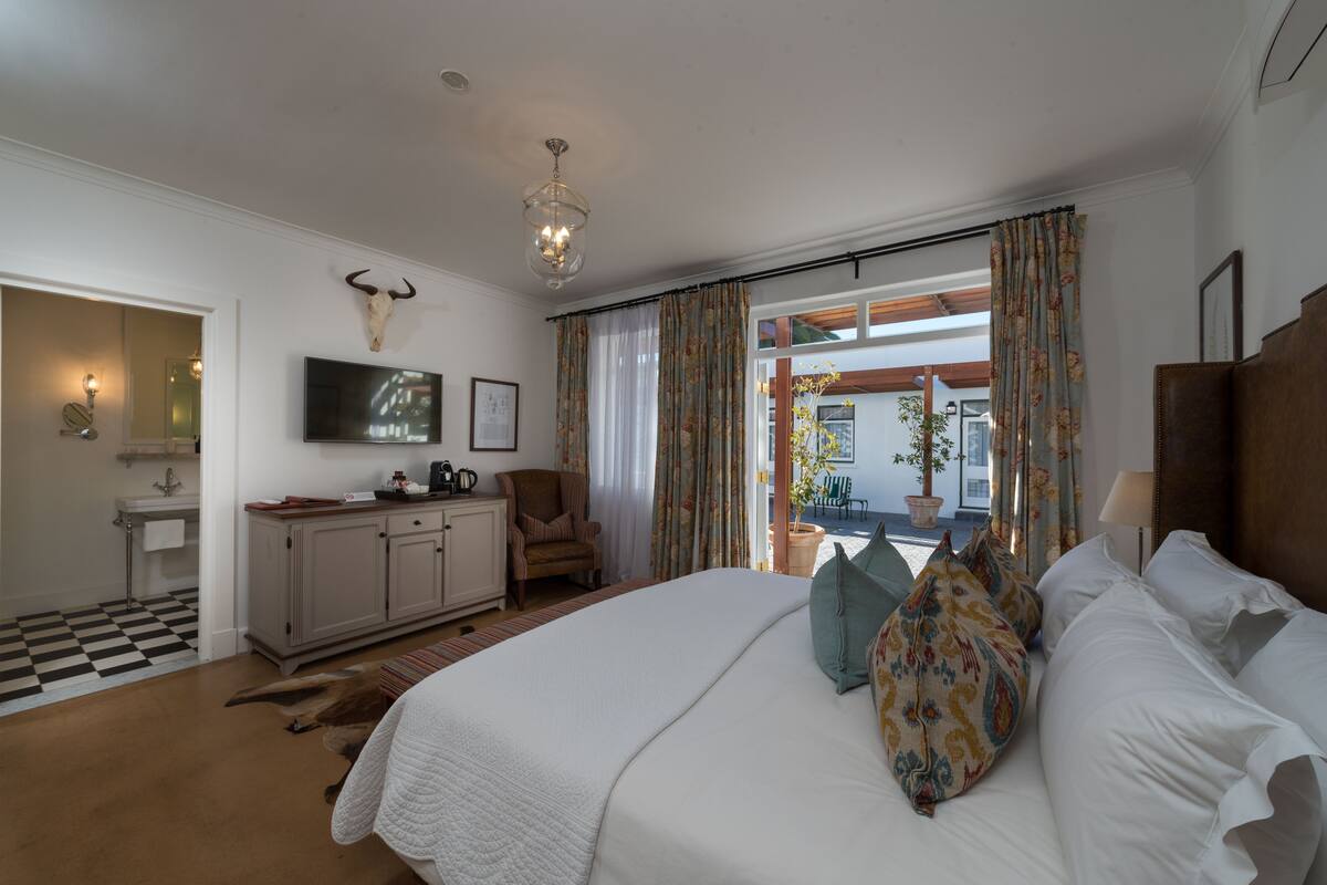 A bedroom with a large bed, a television, a desk, and a chair. There is a window with curtains and a view of the outside.