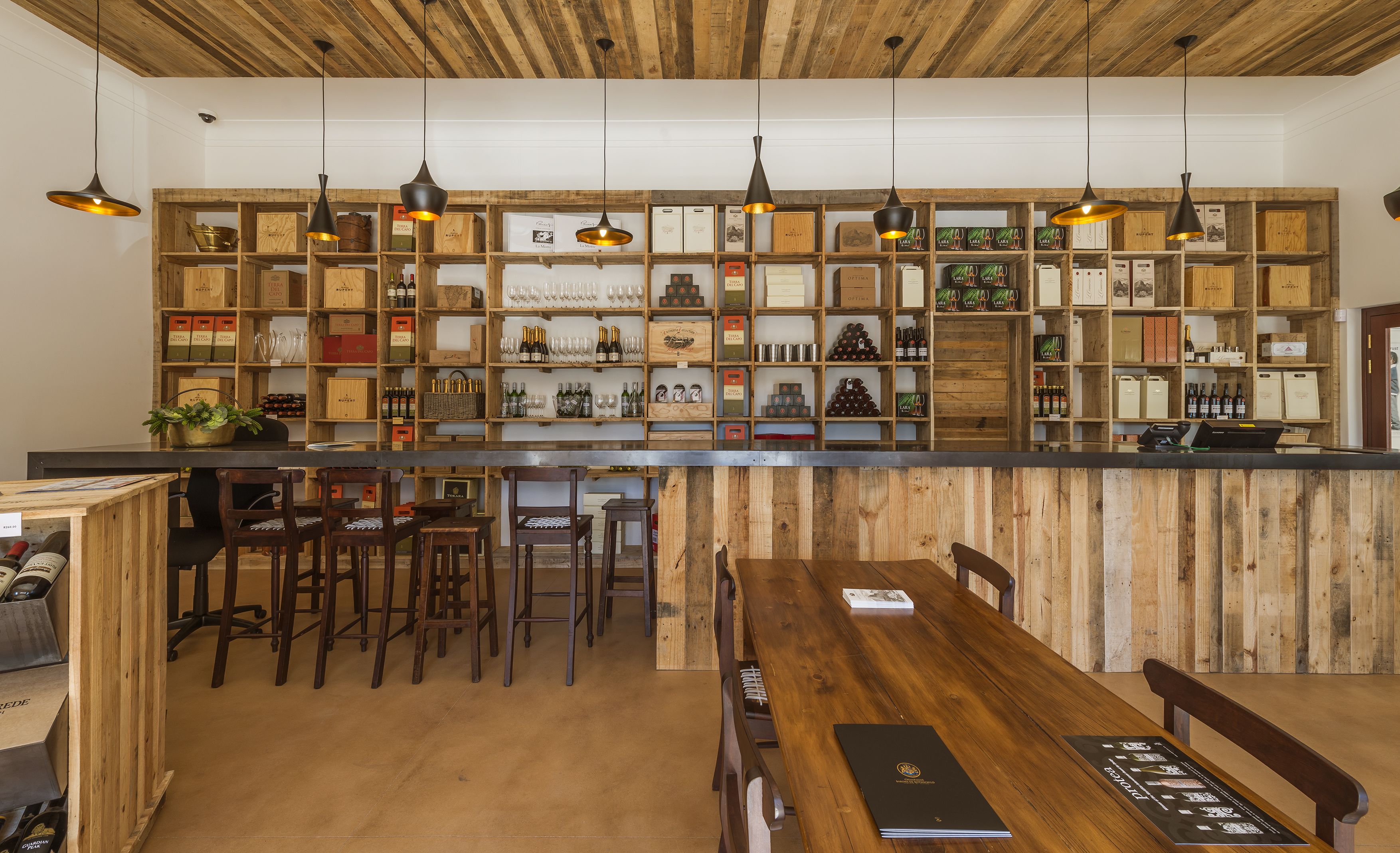 A bar area with wooden shelves displaying bottles and glasses, wooden chairs, and a wooden counter.