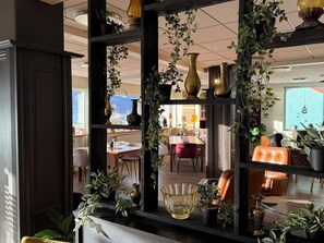 Una habitación con una estantería de madera oscura, varias plantas en macetas y vistas a un comedor con mesas y sillas.