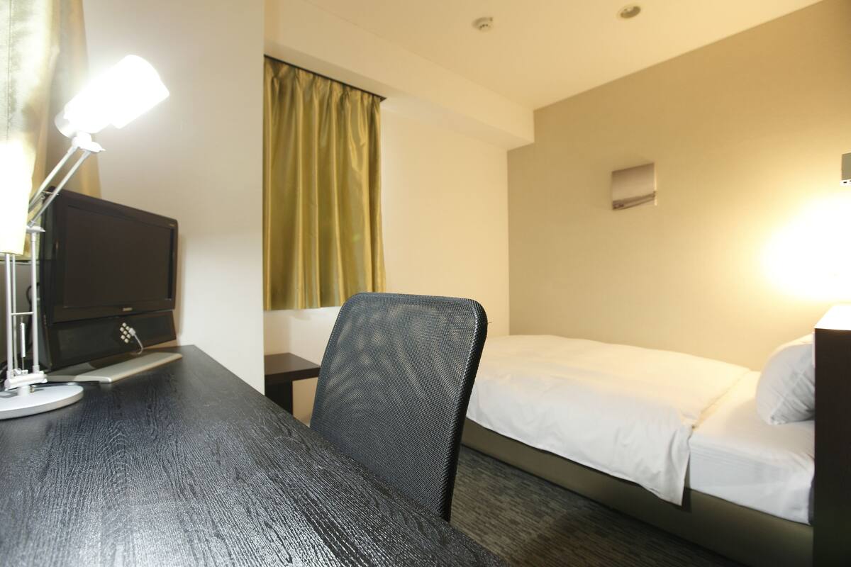 A hotel room with a desk, a chair, a bed, and a window with curtains.