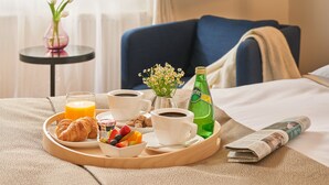 Ett frukostbricka med croissanter, frukt, juice, kaffe och en flaska vatten, placerat på en säng tillsammans med en tidning och en vas med blommor.