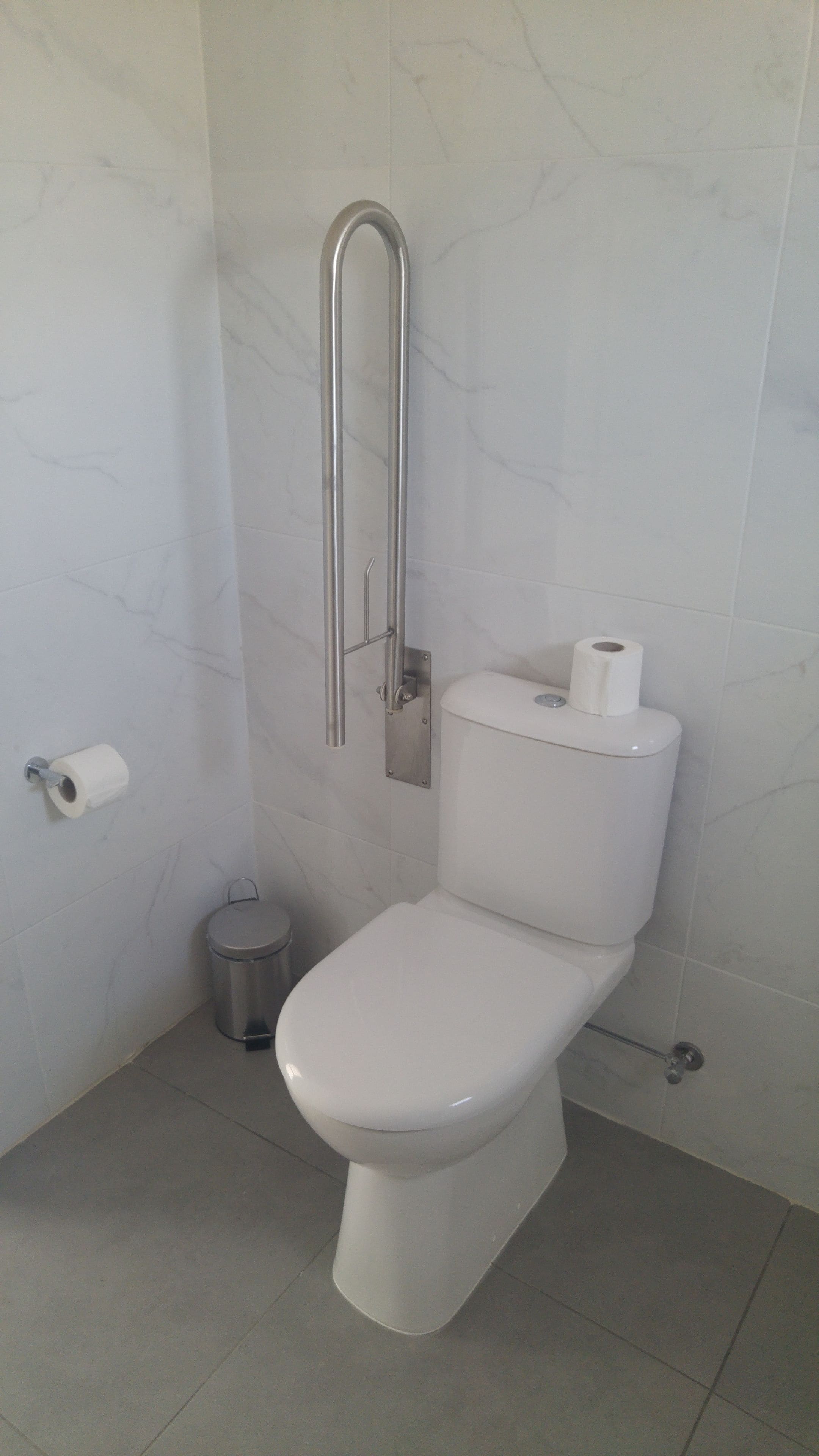 Un baño moderno con inodoro blanco, barra de apoyo, cubo de basura y dispensador de papel higiénico.