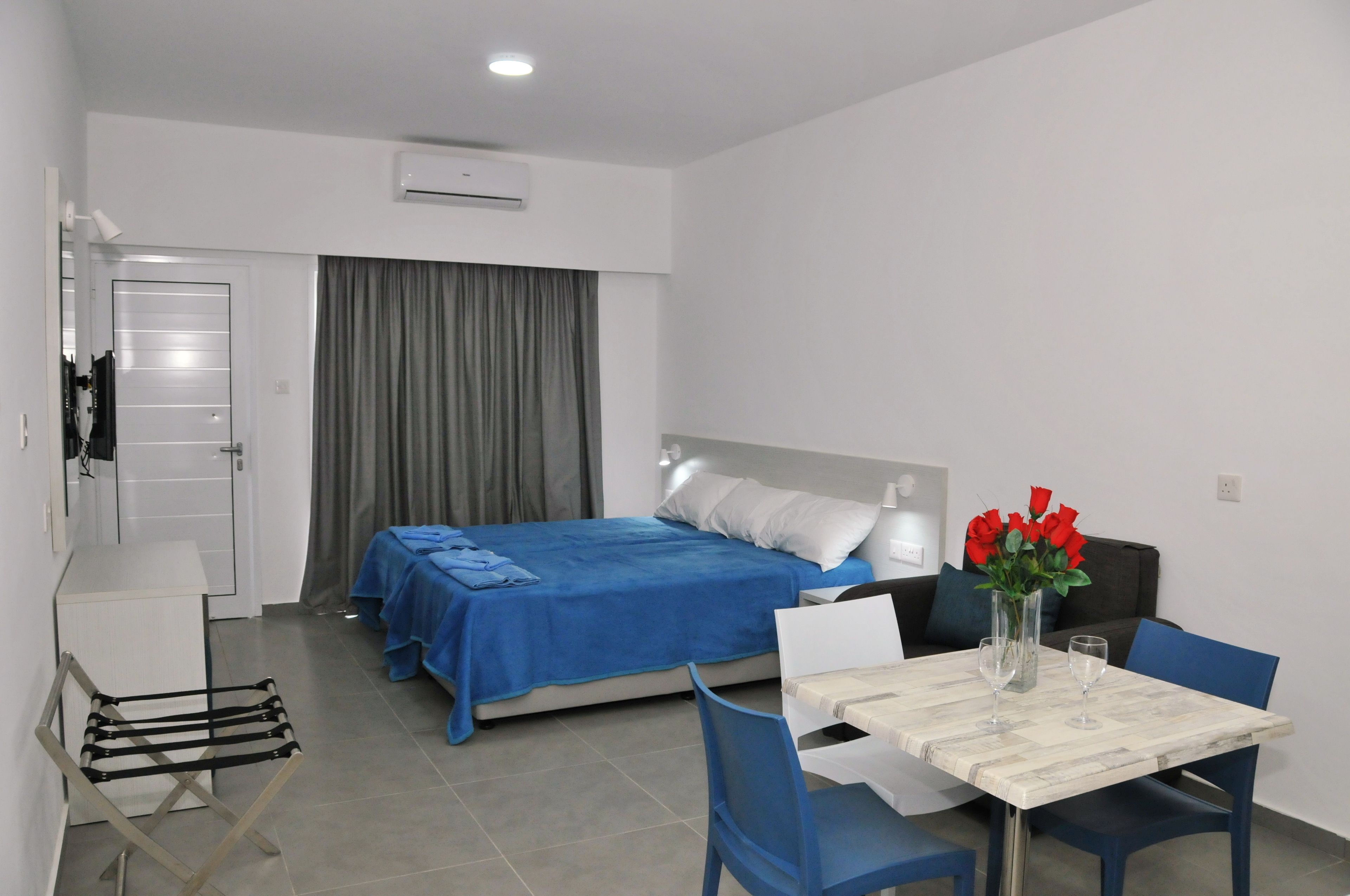 A compact hotel room with a bed, a small table with chairs, a desk, and a chair. There is a window with curtains, an air conditioning unit on the wall, and a coat rack.