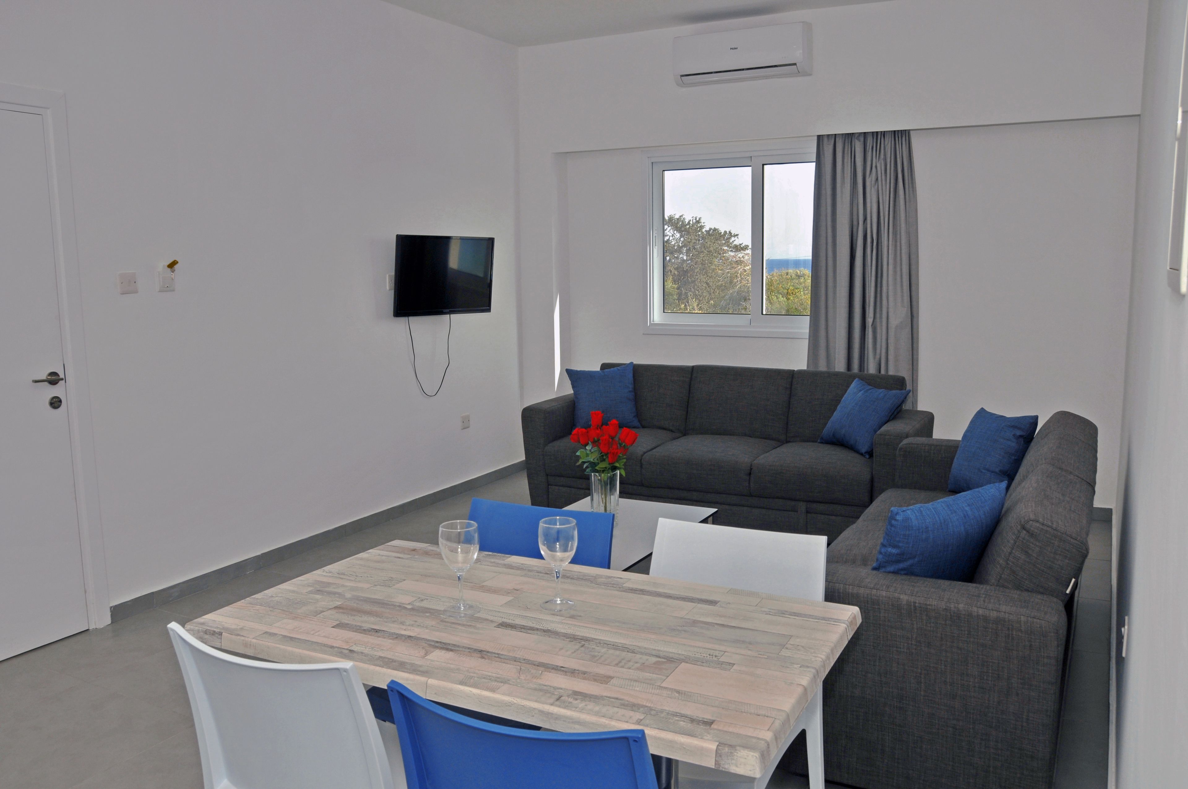 Un salón moderno con un sofá gris, cojines azules, una mesa de madera con dos sillas y un televisor montado en la pared.