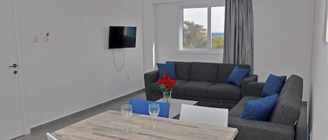 Un salón moderno con un sofá gris, cojines azules, una mesa de madera con dos sillas y un televisor montado en la pared.