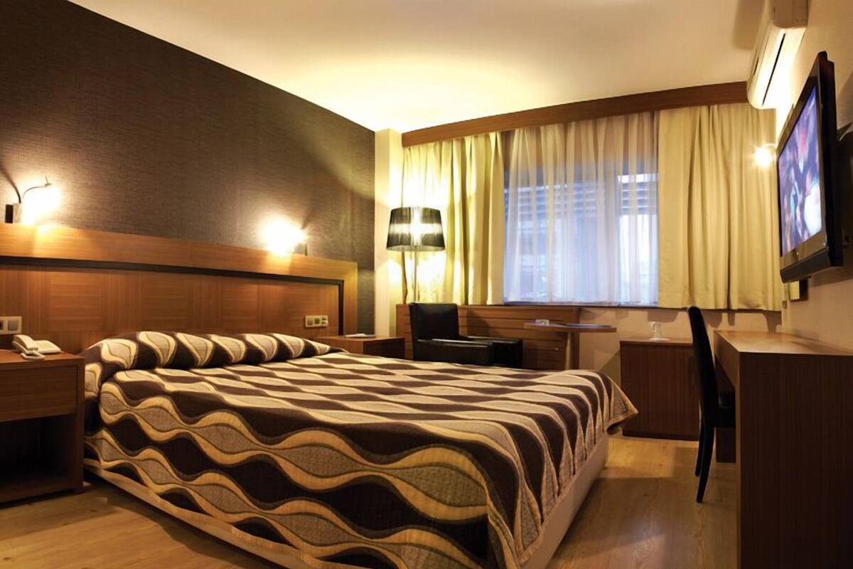 A hotel room with a large bed, a desk, a chair, a TV, and a window with curtains.
