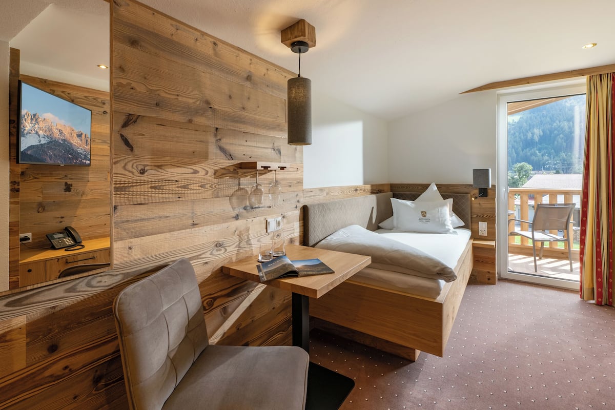 Une chambre d’hôtel avec un mur en bois, un lit avec des oreillers, un bureau avec une chaise, un téléviseur fixé au mur et un balcon offrant une vue sur les montagnes.