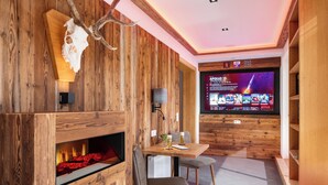 Een gezellige kamer met een open haard, houten wanden, een televisie waarop een film draait, en een tafel met stoelen.