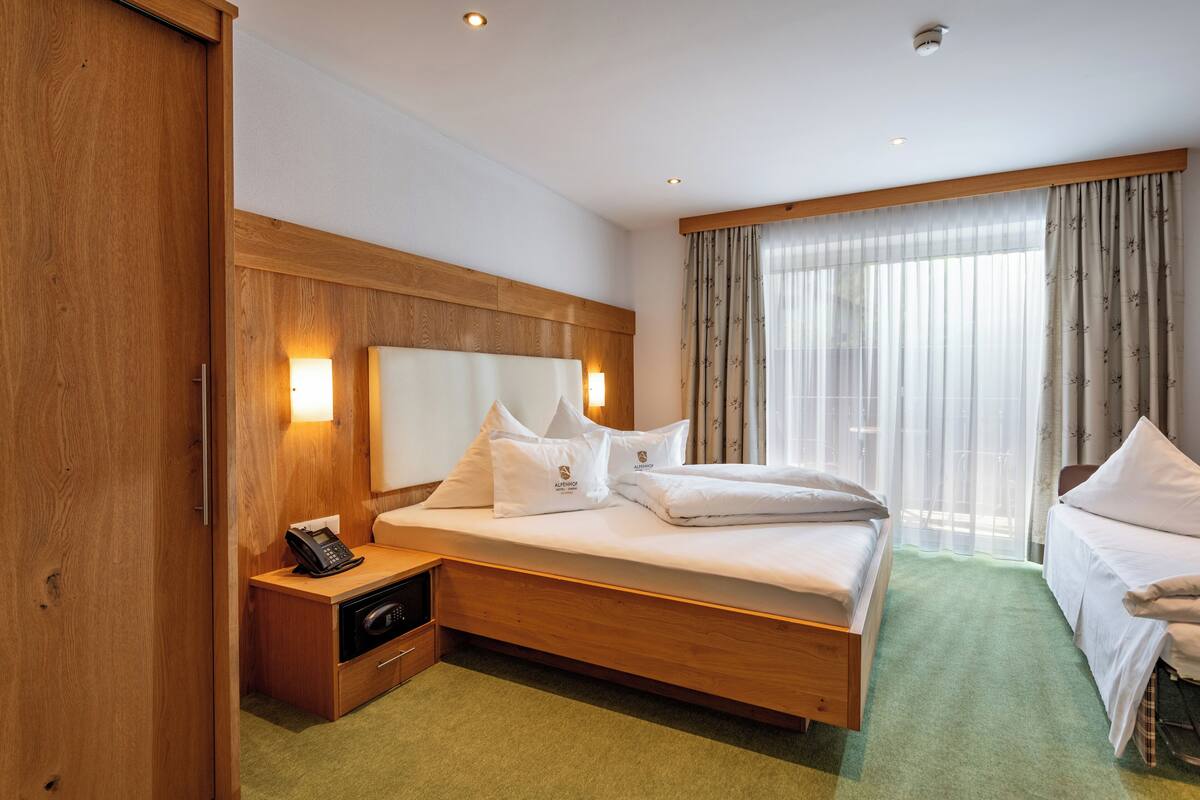 Une chambre d’hôtel avec une tête de lit en bois, un lit recouvert de draps blancs, une table de chevet avec un téléphone, une moquette verte et une fenêtre ornée de rideaux légers.