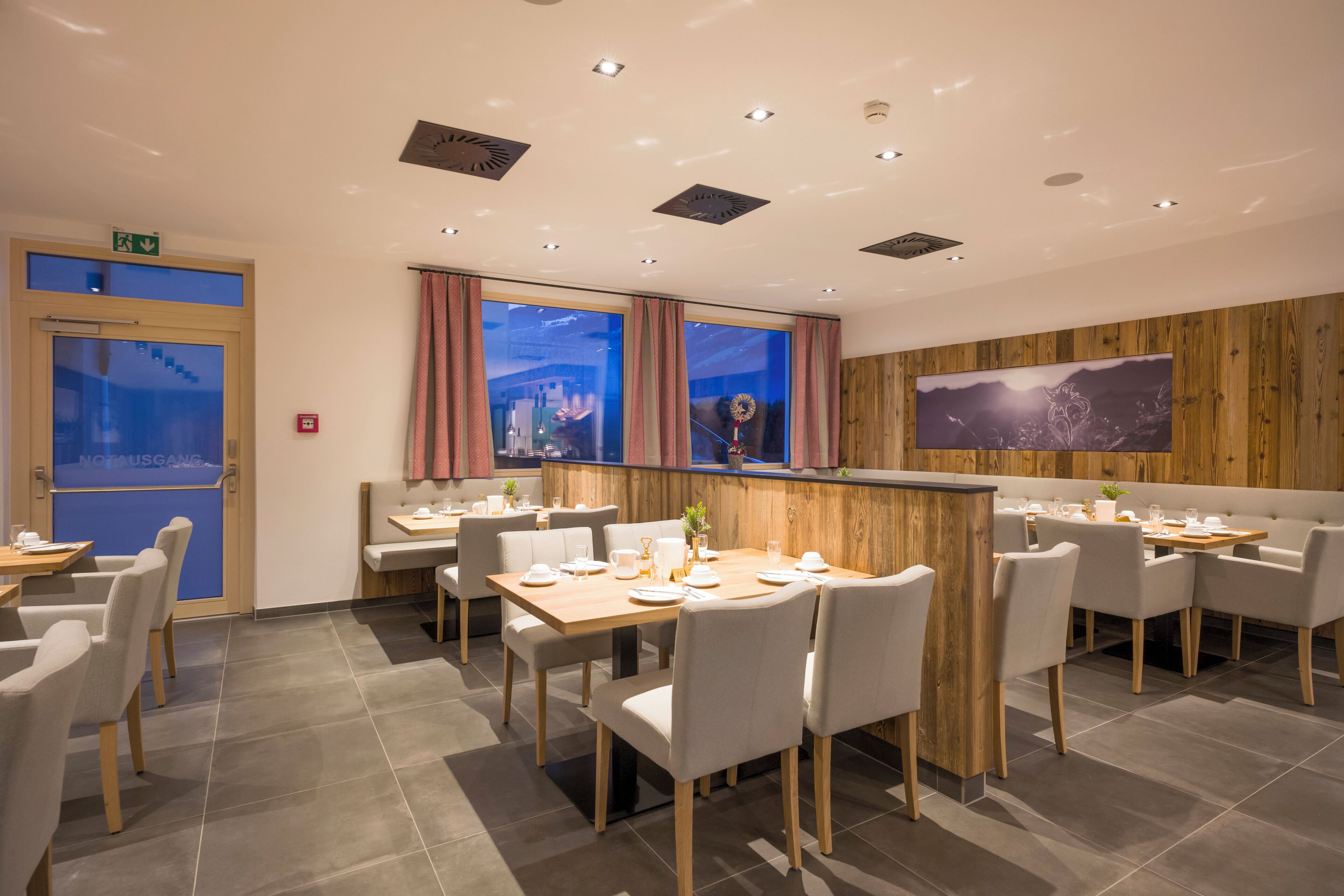 A modern restaurant with multiple dining tables and chairs, a wooden accent wall, and a large TV screen.